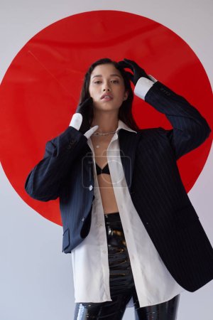 fashion forward, young asian model in bra, white shirt and blazer posing in gloves and latex shorts near red round shaped glass, looking at camera on grey background, personal style, youth 