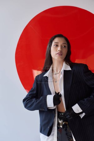 Photo for Fashion photography, young asian model in white shirt and blazer posing in gloves and latex shorts near red round shaped glass, grey background, looking away, personal style, youth trend - Royalty Free Image