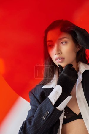 modern woman, fashion photography, young asian model in white shirt and blazer posing in gloves near red round shaped glass, grey background, looking away, personal style, youth trend