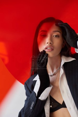 Foto de Moderno individuo, fotografía de moda, joven modelo asiático en camisa blanca y chaqueta posando en guantes cerca de vidrio redondo rojo, fondo gris, cara conmovedora, estilo personal, tendencia juvenil - Imagen libre de derechos