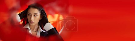 Foto de Moda adelante, joven modelo asiático en camisa blanca y chaqueta posando en guantes detrás de cristal rojo, estilo personal, tendencia juvenil, conceptual, cuello conmovedor y posando en estudio, bandera - Imagen libre de derechos