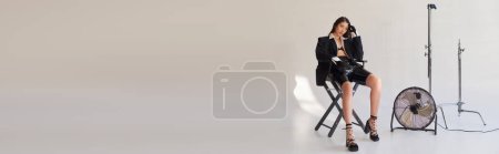 Foto de Fotografía de estudio, mujer asiática joven en blazer, camisa blanca y pantalones cortos de látex sentado en silla plegable cerca de ventilador eléctrico sobre fondo gris, declaración de moda, mirando hacia otro lado, longitud completa, pancarta - Imagen libre de derechos