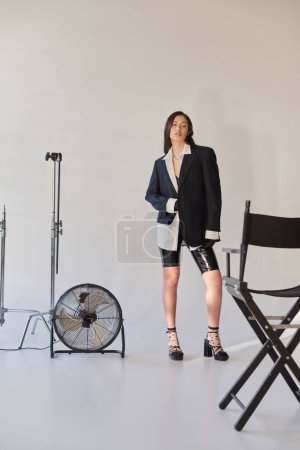 Modestatement, Studiofotografie, junge asiatische Frau in stylischem Look posiert auf grauem Hintergrund, Blazer, weißes Hemd und Latex-Shorts, neben elektrischem Ventilator stehend, persönlicher Stil, volle Länge 