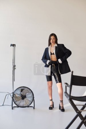 Mode vorne, Studioaufnahmen, junge asiatische Frau in stylischem Look posiert auf grauem Hintergrund, Blazer, weißes Hemd und Latex-Shorts, neben elektrischem Ventilator stehend, konzeptionell, in voller Länge 