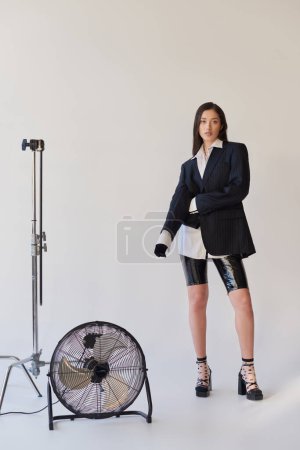 fashion forward, studio photography, young asian woman in stylish look posing on grey background, blazer, white shirt and latex shorts, standing near electric fan, personal style, full length 