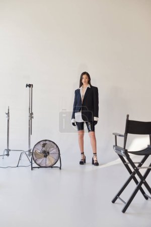 fashion and style, studio photography, young asian woman in stylish look posing on grey background, blazer, white shirt and latex shorts, standing near electric fan, personal style, full length 