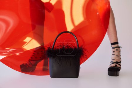 cropped view of woman in dotted socks and high heels standing near round shaped glass on grey background, feathered handbag, female legs, fashion statement, personal style, youth 