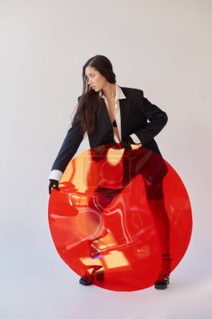 Mode und Stil, Studiofotografie, junge asiatische Frau im stylischen Look posiert in der Nähe von rotem Rundglas, grauem Hintergrund, Blazer und Latex-Shorts, persönlicher Stil, volle Länge 