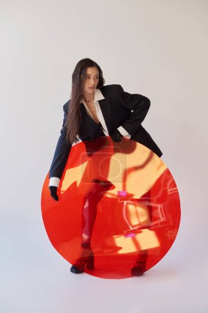 fashion and style, studio photography, young asian model in trendy clothes posing near red round shaped glass, grey background, blazer and latex shorts, personal style, full length 