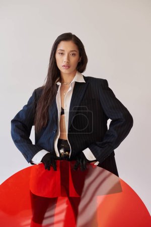 Photo for Edgy style, studio photography, young asian woman in stylish look posing near red round shaped glass, grey background, blazer and latex shorts, youthful fashion, cool style - Royalty Free Image