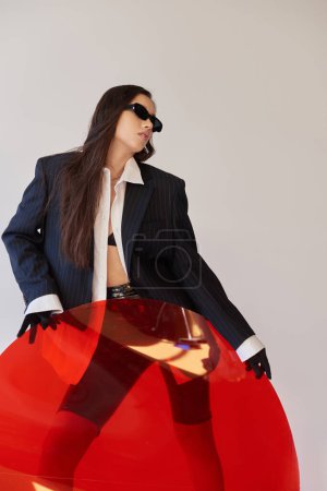 moda moderna, fotografía de estudio, mujer asiática joven en aspecto elegante y gafas de sol posando cerca de vidrio redondo rojo, fondo gris, blazer y pantalones cortos de látex, moda juvenil, estilo fresco 