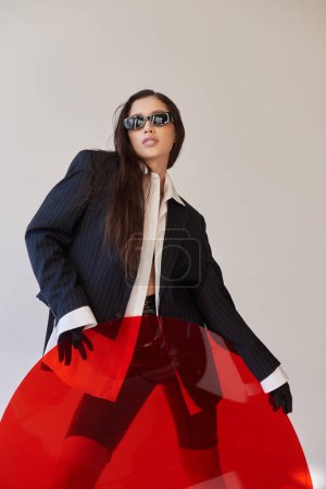 Photo for Edgy style, studio photography, young asian model in stylish look and sunglasses posing near red round shaped glass, grey background, blazer and latex shorts, youthful fashion, modern woman - Royalty Free Image