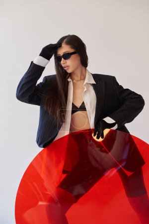 cool style, studio photography, young asian model in stylish look and sunglasses posing near red round shaped glass, grey background, blazer and latex shorts, youthful fashion, modern woman 