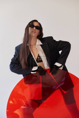 young asian model in stylish look and sunglasses posing near red round shaped glass, grey background, blazer and latex shorts, youthful fashion, modern woman, edgy style, studio photography 