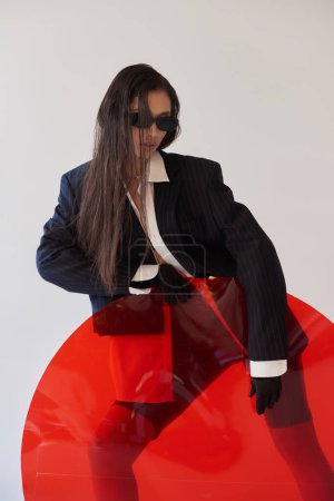 Foto de Moda de látex, modelo asiático joven en aspecto elegante y gafas de sol posando cerca de vidrio redondo rojo, fondo gris, blazer y pantalones cortos de látex, moda juvenil, mujer moderna, fotografía de estudio - Imagen libre de derechos