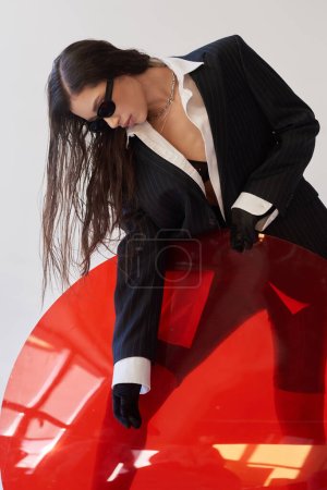 Foto de Modelo asiático atractivo en aspecto elegante y gafas de sol posando cerca de vidrio redondo rojo en forma, fondo gris, chaqueta y pantalones cortos de látex, mujer joven y moderna, estilo vanguardista, fotografía de estudio - Imagen libre de derechos