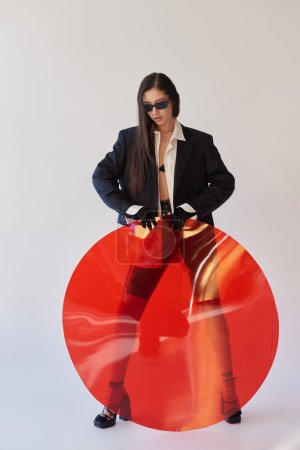 attraktives asiatisches Model in stylischem Look und Sonnenbrille posiert mit rotem Rundglas, grauem Hintergrund, Blazer und Latex-Shorts, jugendliche und moderne Frau, Mode nach vorne, Studiofotografie 