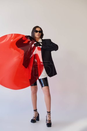pretty asian model in stylish look and sunglasses posing holding red round shaped glass, grey background, blazer and latex shorts, youthful and modern woman, fashion statement, studio photography 
