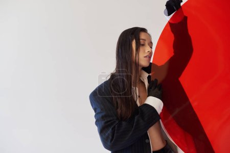 pretty asian woman in trendy outfit holding red round shaped glass, grey background, blazer and black latex shorts, youthful model in gloves, fashion choices, studio photography, conceptual 