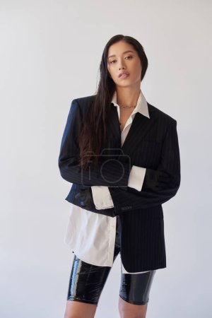 moda adelante, joven mujer asiática en traje de moda con pantalones cortos de látex posando sobre fondo gris, chaqueta y guantes negros, de pie con los brazos cruzados, modelo juvenil, fotografía de estudio, conceptual 