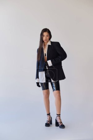 estilo fresco, mujer asiática joven en traje de moda con pantalones cortos de látex posando sobre fondo gris, chaqueta y guantes negros, modelo juvenil en tacones altos, fotografía de estudio, conceptual, longitud completa