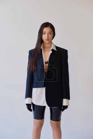 jeune mode, femme asiatique brune en short en latex posant sur fond gris, chemise blanche, blazer et gants noirs, jeune mannequin, studio photographie, conceptuel 
