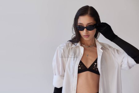 bra look, fashion statement, stylish asian woman in sunglasses posing on grey background, young model in black gloves and white shirt, perfect skin, conceptual, trendy outfit, youthful  