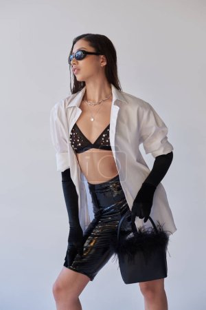 personal style, fashion forward, asian woman in sunglasses posing with feathered purse on grey background, young model in latex shorts, black gloves and white shirt, conceptual 