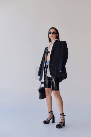 trendy look, fashion statement, brunette asian woman in sunglasses posing with feathered purse on grey background, model in latex shorts, bra, black jacket and gloves, youth, full length 