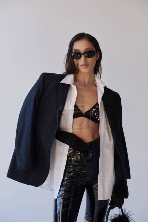 persönlicher Stil, brünette asiatische Frau mit dunkler Sonnenbrille posiert mit gefiederter Handtasche auf grauem Hintergrund, junges Model in Latexhosen, BH, Blazer und schwarzen Handschuhen, Jugend und Stil 