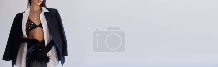 persönlicher Stil, brünette Frau im BH posiert mit gefiederter Handtasche auf grauem Hintergrund, junges Model in Latex-Shorts, Blazer und schwarzen Handschuhen, Jugend und Stil, abgeschnittenes Image, Banner