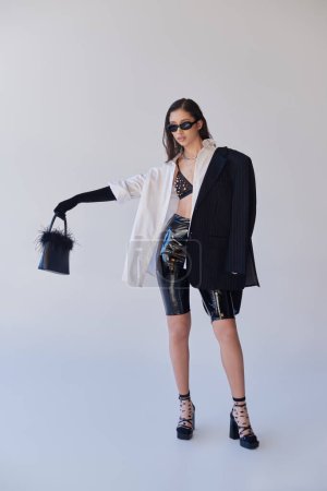 edgy style, Studiofotografie, junge asiatische Frau in stylischem Look und Sonnenbrille posiert mit gefiederter Handtasche auf grauem Hintergrund, Blazer und Latex-Shorts, jugendliche Mode, volle Länge 