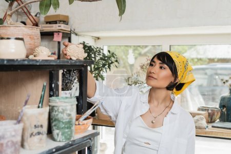 Junge asiatische Künstlerin in Arbeitskleidung und Kopftuch nimmt Tonerzeugnisse aus dem Regal, während sie neben Töpferwerkzeugen im Töpferkurs steht, Töpferatelier mit Kunsthandwerker bei der Arbeit