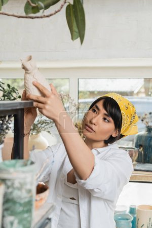 Junge asiatische Künstlerin in Kopftuch und Arbeitskleidung nimmt Tonerzeugnis aus dem Regal, während sie im verschwommenen Töpferkurs steht und arbeitet, Töpferatelier mit Kunsthandwerker bei der Arbeit