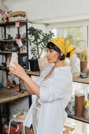 Junge brünette asiatische Künstlerin in Kopftuch und Arbeitskleidung, die Tonerzeugnis hält, während sie im verschwommenen Töpferkurs im Hintergrund steht, Töpferatelier mit Kunsthandwerker bei der Arbeit