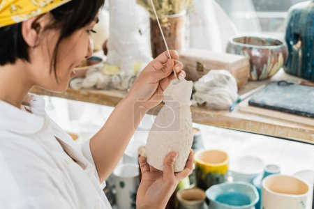 Téléchargez les photos : Vue latérale d'une jeune artiste asiatique souriante et floue tenant un bâton en bois et des produits en argile tout en travaillant dans une classe de poterie floue en arrière-plan, un atelier de poterie avec un artisan au travail - en image libre de droit