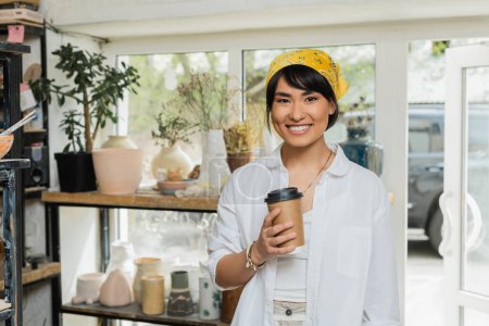 Photo for Positive young asian artist in headscarf and workwear looking at camera and holding coffee to go while standing in blurred pottery class at background, pottery workshop with skilled artisan - Royalty Free Image