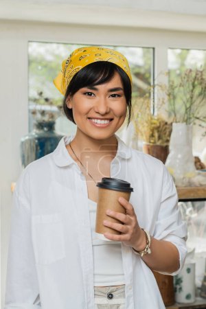 Portrait of smiling young asian female artisan in headscarf and workwear looking at camera and holding coffee to go while standing in blurred pottery class, pottery workshop with skilled artisan