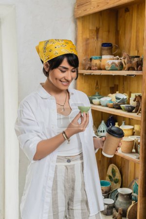 Cheerful young asian artist in headscarf and workwear holding coffee to go and clay product and standing near shelves with sculptures at background, pottery workshop with skilled artisan