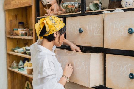 Photo for Side view of young asian female artist in headscarf and workwear opening cupboard while working in blurred pottery workshop, pottery workshop with skilled artisan - Royalty Free Image