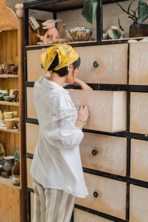 Junge asiatische Keramikerin mit Kopftuch und Arbeitskleidung öffnet Schrank während der Arbeit und steht in der Nähe von Tonerzeugnissen auf Regalen im Keramikatelier, Töpferwerkstatt mit qualifizierter Kunsthandwerkerin