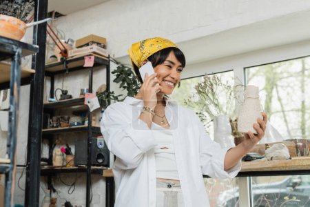 Fröhliche junge asiatische Kunsthandwerkerin in Kopftuch und Arbeitskleidung, die mit dem Smartphone spricht und Tonskulpturen hält, während sie in der Keramikwerkstatt steht, Kunsthandwerkerin im Töpferatelier, die sich auf die Schöpfung konzentriert