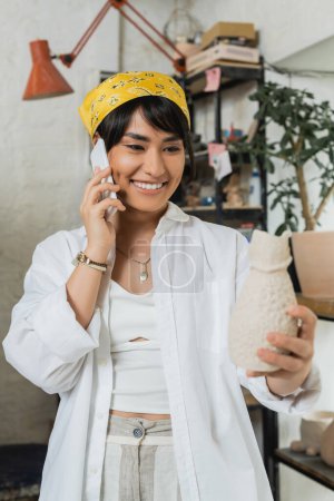 Positive young asian craftswoman in headscarf and workwear talking on smartphone and holding clay sculpture while standing in ceramic workshop, artisan in pottery studio focusing on creation