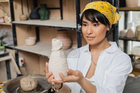 Smiling young asian artist in headscarf and workwear holding and looking at clay sculpture while working in ceramic workshop, artisan in pottery studio focusing on creation