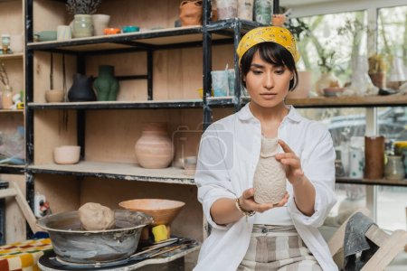 Young asian brunette female potter in headscarf and workwear holding clay sculpture near pottery wheel while working in ceramic workshop, craftsmanship in pottery making