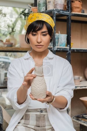 Portrait of young asian female potter in headscarf and workwear holding clay sculpture and working in blurred ceramic workshop, craftsmanship in pottery making