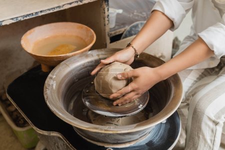 Cropped view of young female artisan in workwear molding clay and working with pottery wheel near bowl with water in ceramic workshop, craftsmanship in pottery making