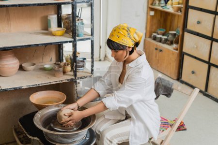 Junge asiatische Kunsthandwerkerin mit Kopftuch und Arbeitskleidung, die Ton auf Töpferscheibe formt und in der Nähe von Töpferwerkzeugen in einer verschwommenen Keramikwerkstatt arbeitet, Kunsthandwerk in der Töpferei