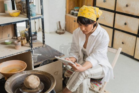 Junge brünette asiatische Kunsthandwerkerin in Kopftuch und Arbeitskleidung mit digitalem Tablet, während sie in der Nähe der Töpferscheibe in einer verschwommenen Keramikwerkstatt sitzt, Handwerkskunst in der Töpferei