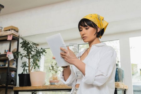 Young brunette asian female artist in headscarf and workwear using digital tablet while working in blurred ceramic workshop at background, craftsmanship in pottery making
